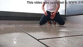 Masked figure masturbates outdoors in wet jeans on a rainy balcony.