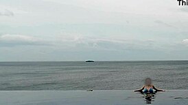 brunette asian student with ponytail and small waist relaxes by infinity pool in swimsuit